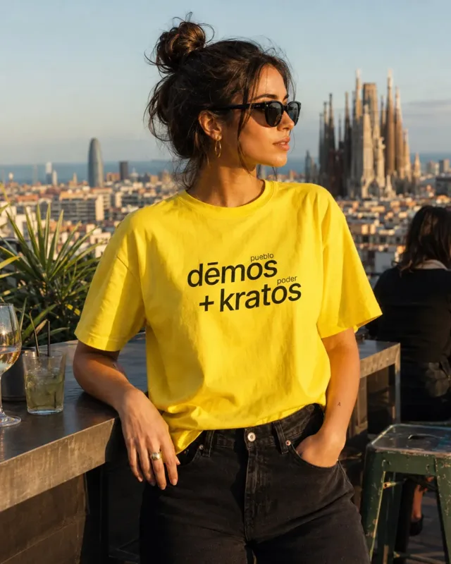 Modelo con camiseta amarilla democracia21 con el mensaje dēmos + kratos, etiquetada "pueblo" y "poder", en una azotea con la Sagrada Família al fondo